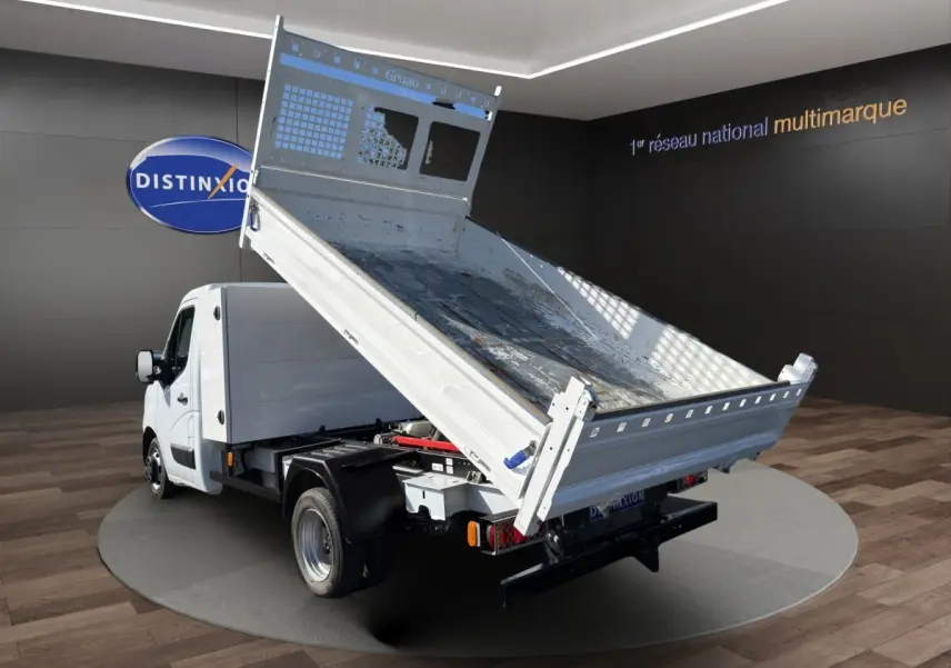 Renault Master blanc en 3/4 arrière avec benne basculante relevée, sur fond intérieur showroom.