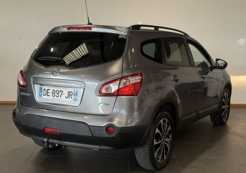 Vue arrière droite d'un Nissan Qashqai+2 gris 2014 avec feux arrière rouges et attelage de remorque visible.