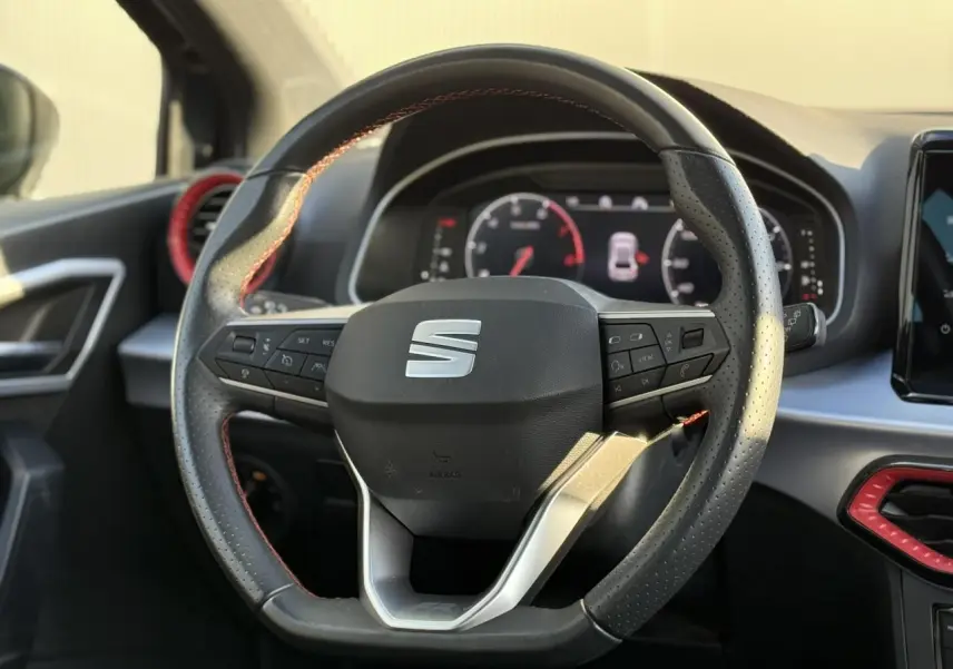 Vue rapprochée du volant perforé noir avec logo SEAT, entouré d'inserts rouges et tableau de bord numérique en arrière-plan.