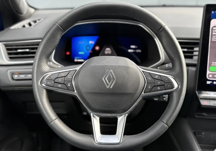 Vue rapprochée du volant noir perforé avec logo Renault, tableau de bord numérique et écran tactile à droite.
