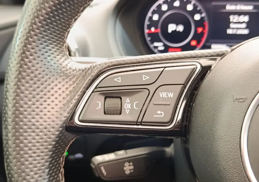 Gros plan sur les commandes gauche du volant en cuir perforé d’un Audi Q2 noir, avec tableau de bord numérique en arrière-plan.