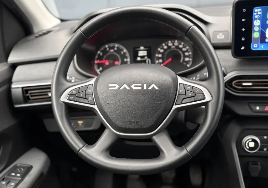 Vue frontale du volant noir Dacia avec commandes intégrées et tableau de bord numérique en arrière-plan.