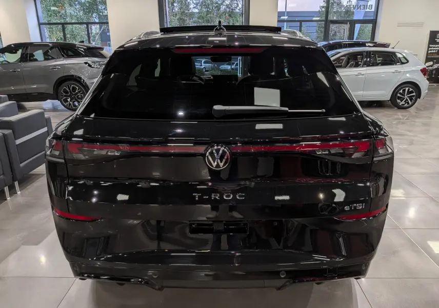 Vue arrière d’un Volkswagen T-Roc eTSI noir Ébène 2026 avec feux LED reliés et logo central lumineux dans un showroom.