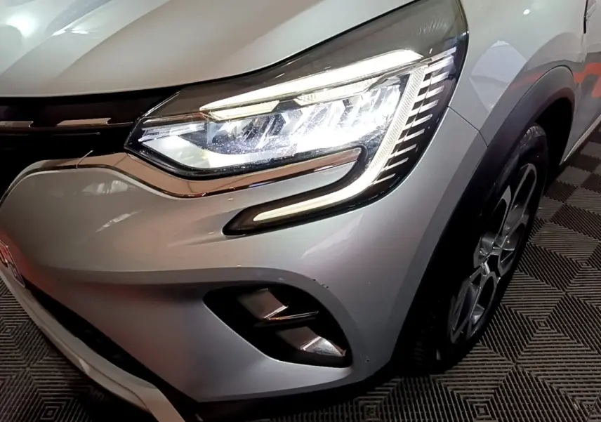 Gros plan sur le phare avant droit allumé d’un Renault Captur gris clair avec jante et partie de la calandre visibles.
