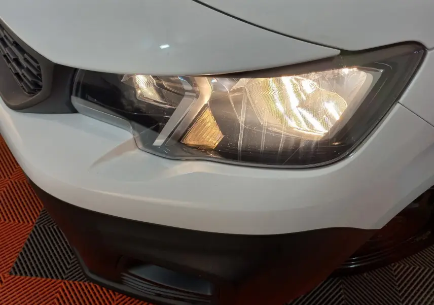 Gros plan sur le phare avant droit allumé d’un Peugeot Partner blanc, avec partie de la calandre et pare-chocs noir visible.