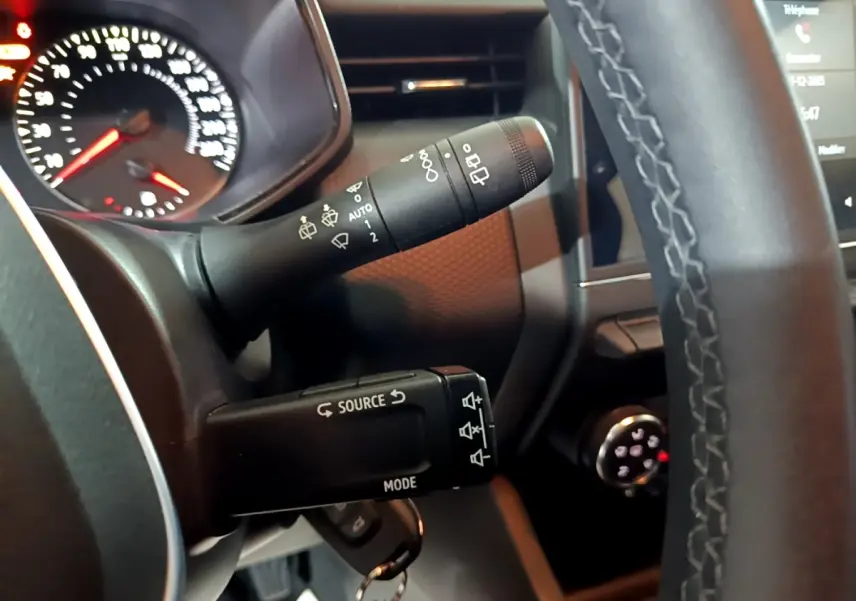 Gros plan sur la colonne de direction et commandes au volant cuir d'une Renault Clio gris clair, tableau de bord partiellement visible.