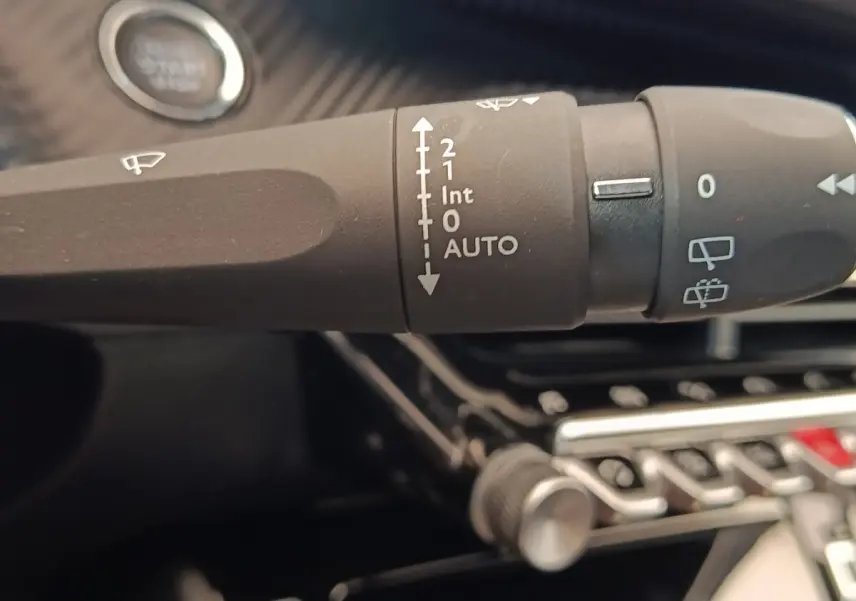 Gros plan sur la commande des essuie-glaces noire dans l'habitacle d'une Peugeot 208 blanche, avec bouton démarrage visible.