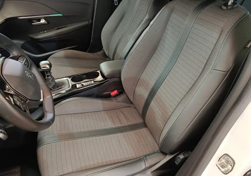 Vue intérieure côté conducteur de la Peugeot 208 blanche, montrant les sièges gris tissu avec surpiqûres et volant cuir multifonctions.