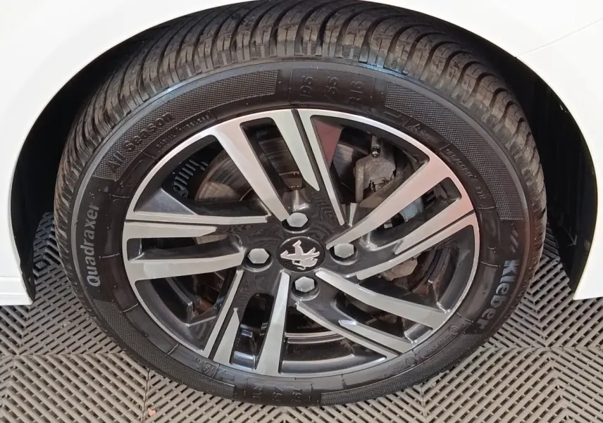 Gros plan sur la roue avant droite d'une Peugeot 208 blanche, jante alliage noire et argent avec logo Peugeot central.