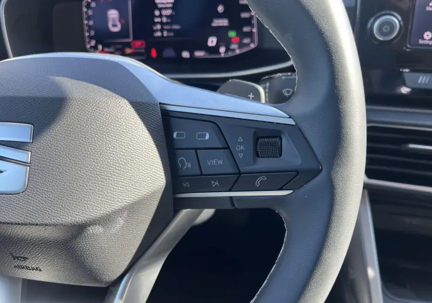 Gros plan sur le volant cuir multifonction de la SEAT LEON IV 2023 avec commandes vocales et écran digital en arrière-plan.