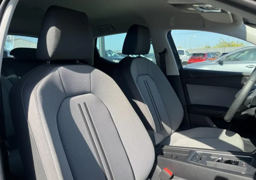 Intérieur avant de la SEAT LEON IV gris Magnetic Metal, sièges tissu gris et accoudoir central visible au soleil.