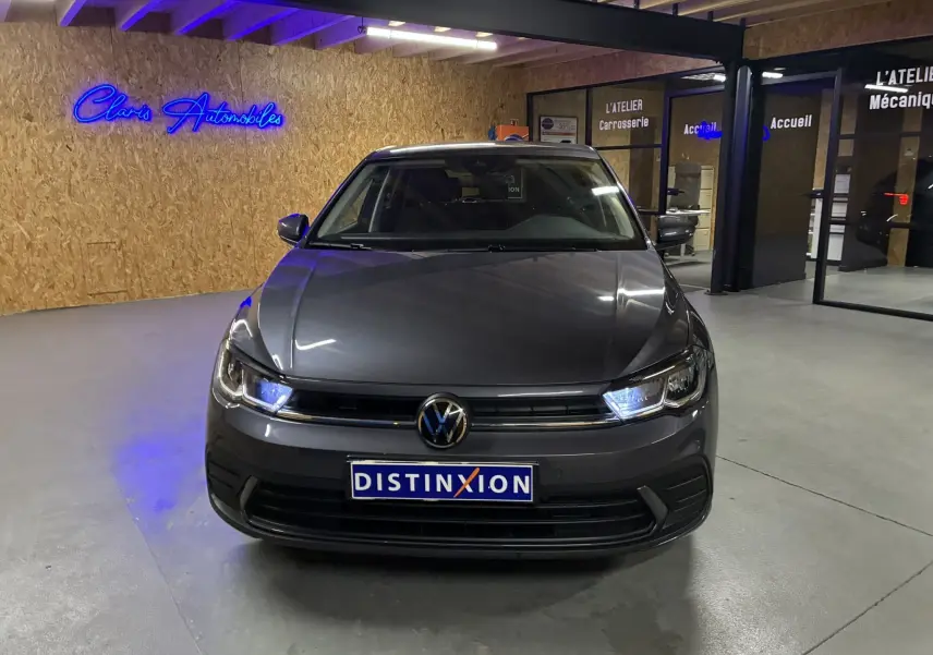 Volkswagen Polo gris vue de face en intérieur, avec phares allumés et plaque Distinxion visible.