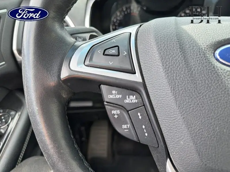 Gros plan sur les commandes à gauche du volant cuir noir du Ford Edge gris Ingot, avec boutons de régulateur et menu.