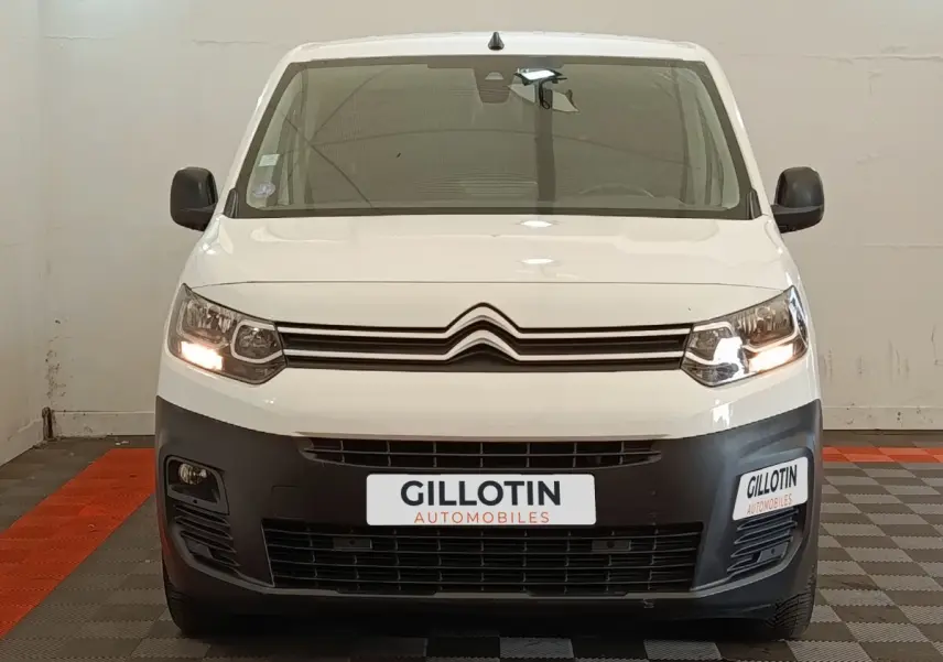 Vue avant d'un Citroën Berlingo blanc avec calandre noire et phares allumés dans un garage.