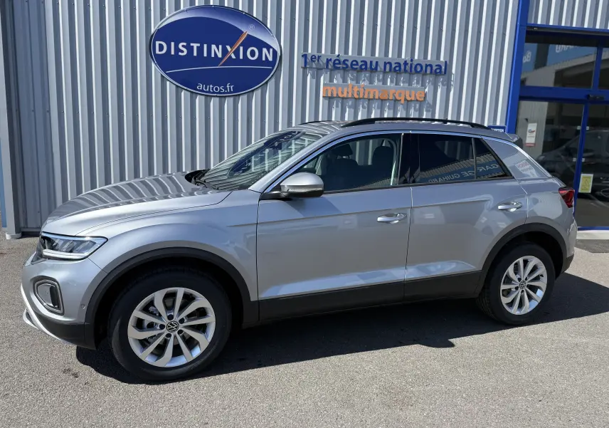 Volkswagen T-Roc 1.5 TSI 150 DSG7 gris pyrite vu de profil côté gauche, stationné devant un bâtiment commercial.