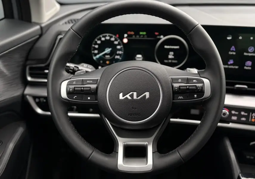 Volant cuir noir du Kia Sportage 2025 avec commandes intégrées, tableau de bord numérique et écran tactile visible en arrière-plan.