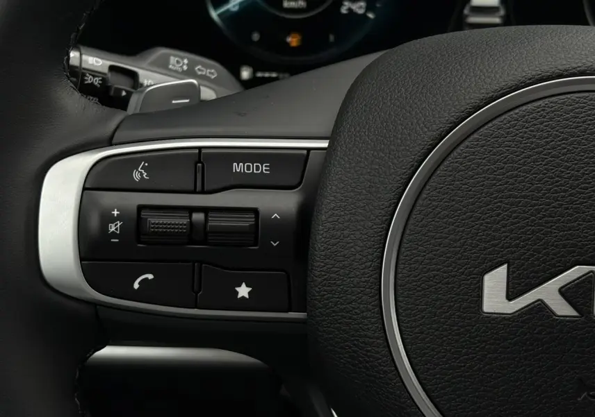 Gros plan sur les commandes gauche du volant cuir noir du Kia Sportage 2025, avec boutons multifonctions et logo central.