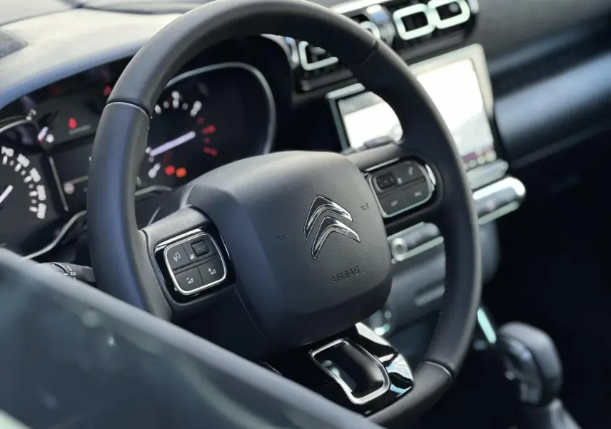Vue rapprochée du volant noir de la Citroën C3 Aircross 2024 avec commandes intégrées et tableau de bord en arrière-plan.