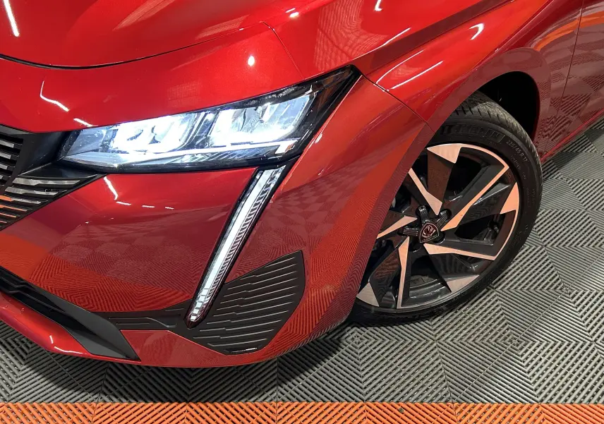 Gros plan sur l'avant droit rouge vif de la Peugeot 308 2024, mettant en valeur le phare LED et la jante alliage noire et argent.