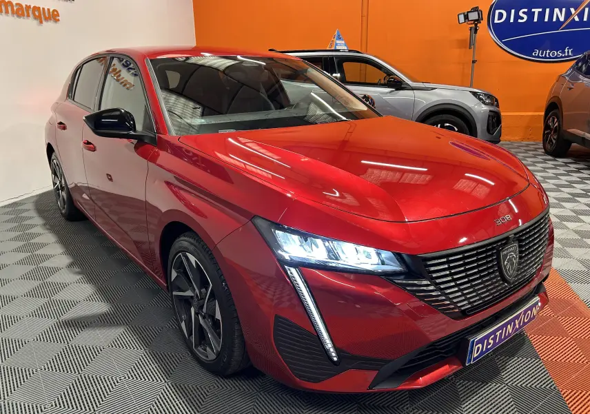 Peugeot 308 rouge vue 3/4 avant droit, phares LED allumés et calandre noire distinctive en showroom.