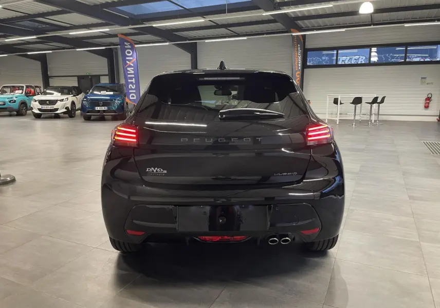 Vue arrière d'une Peugeot 208 noire métallisée avec feux arrière allumés et double sortie d'échappement visible en intérieur showroom.