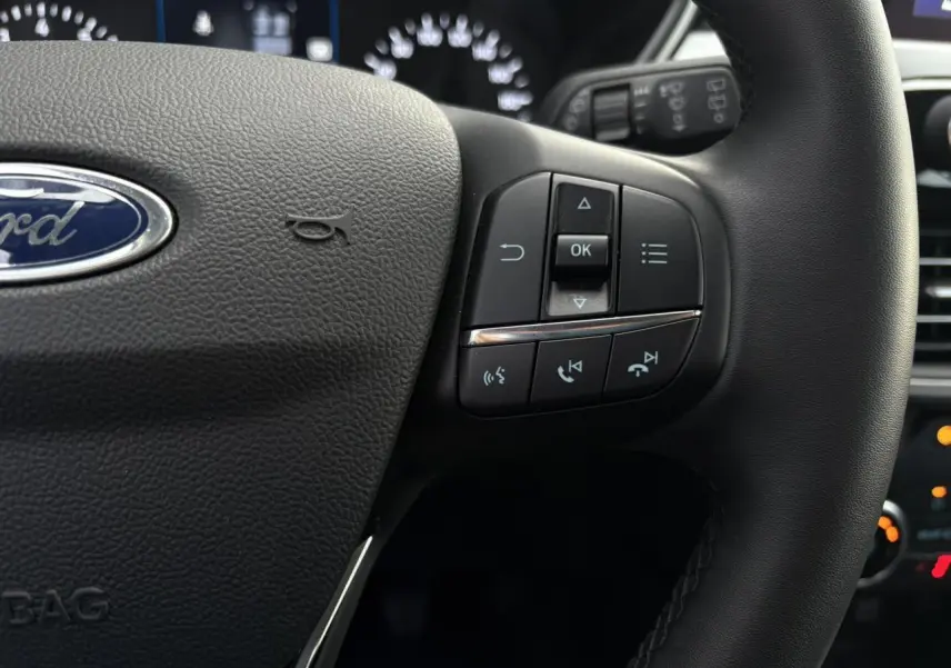 Gros plan sur le volant multifonctions en cuir du Ford Kuga 2024, avec commandes intégrées et logo Ford visible.