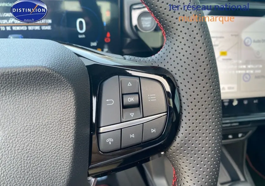 Gros plan sur les commandes du volant perforé avec surpiqûres rouges du Ford Puma rouge, tableau de bord et écran tactile en arrière-plan.