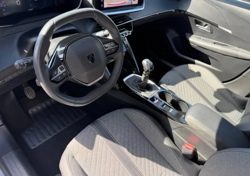 Vue intérieure côté conducteur de la Peugeot 208 1.2 100ch avec volant compact, sellerie grise et boîte manuelle.
