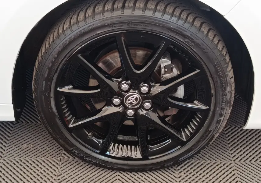 Gros plan sur la roue avant droite noire brillante d'une Toyota Yaris hybride blanche, avec logo Toyota au centre.