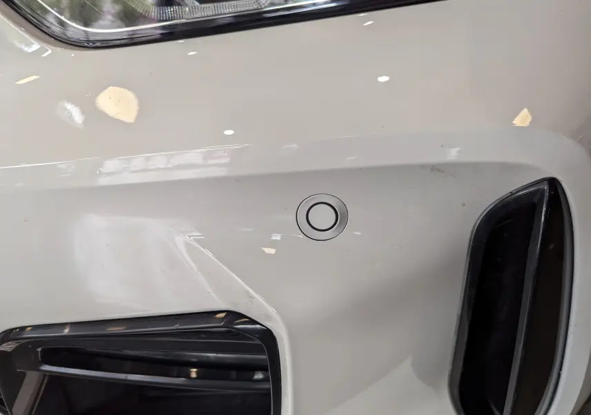 Gros plan sur le pare-chocs avant blanc alpin d'une BMW Série 1 F40 18d M Sport, avec capteur de stationnement visible.