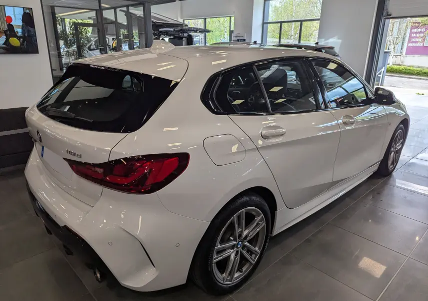 BMW Série 1 F40 18d blanc alpin vue 3/4 arrière droit en intérieur avec jantes alliage et feux LED distinctifs.