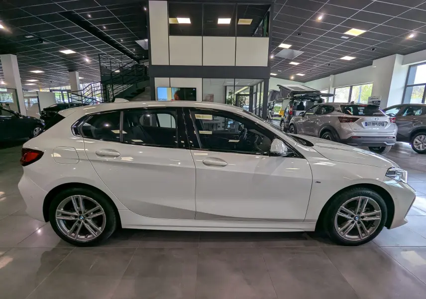 BMW Série 1 F40 18d blanc alpin vue de profil côté gauche en intérieur avec jantes alliage M Sport visibles.