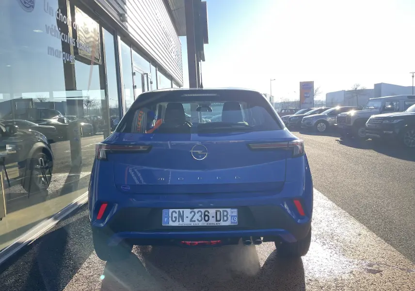 Vue arrière d’un Opel Mokka bleu Voltak 2023 garé devant un bâtiment vitré par une journée ensoleillée.