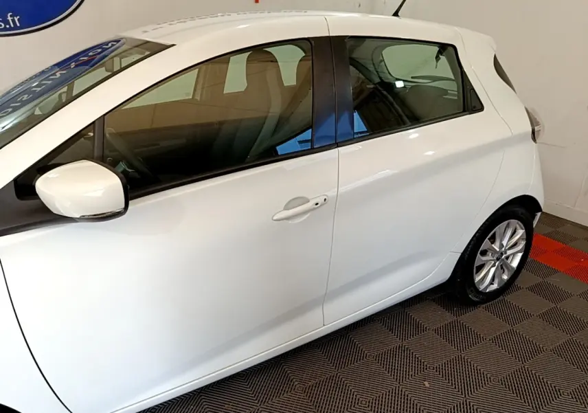 Vue latérale côté gauche d'une Renault ZOE blanche 2019 en intérieur, avec rétroviseur et poignées de porte visibles.