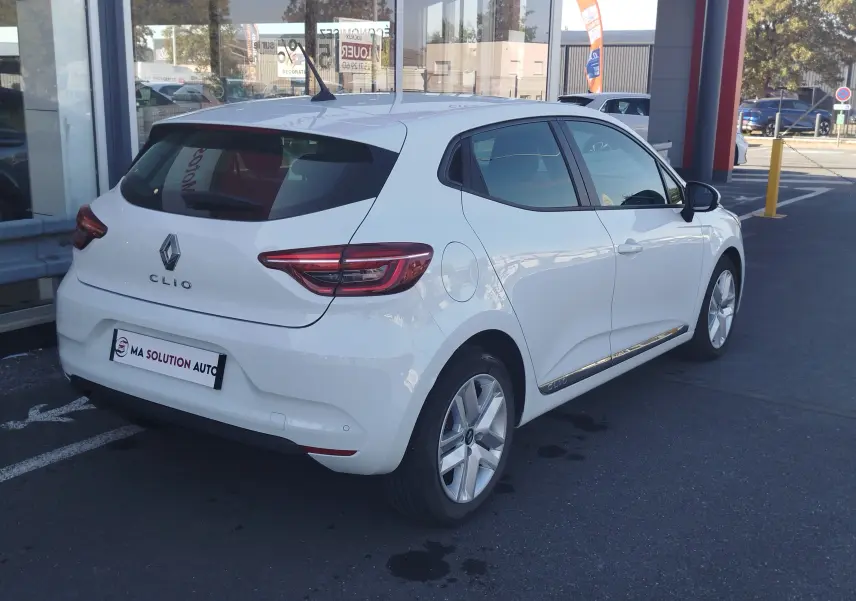 Renault Clio Business blanche vue en 3/4 arrière droit, avec jantes alliage et vitres teintées, stationnée devant un bâtiment.