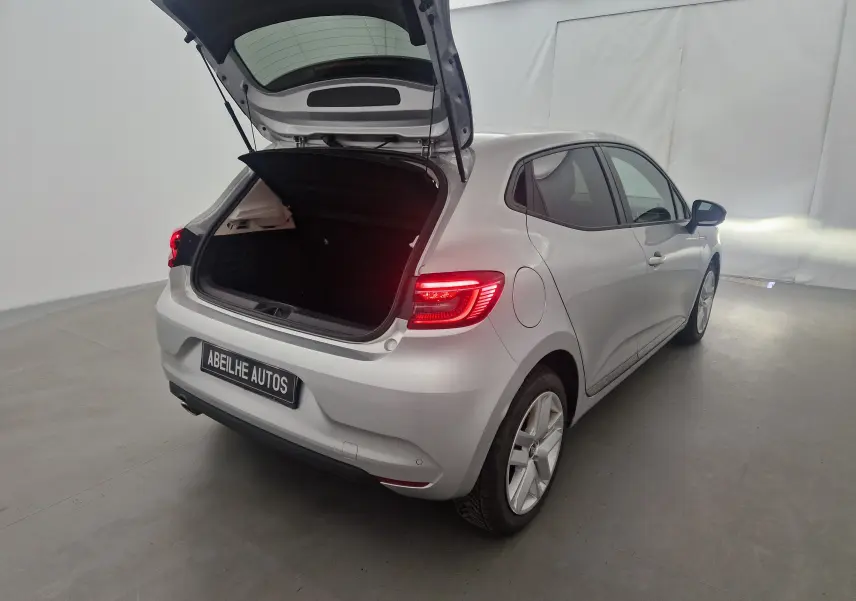 Renault Clio V gris platine vue 3/4 arrière droit avec coffre ouvert et feux arrière allumés dans un studio blanc.