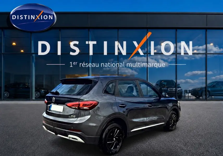 Vue 3/4 arrière droite d’un MG ZS Camden grey hybride 2026 avec feux arrière LED et jantes noires devant un showroom Distinxion.