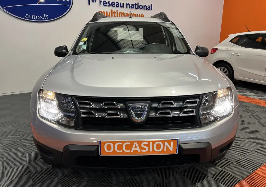 Vue frontale d'un Dacia Duster gris 2017 avec phares allumés et plaque "OCCASION" en concession.
