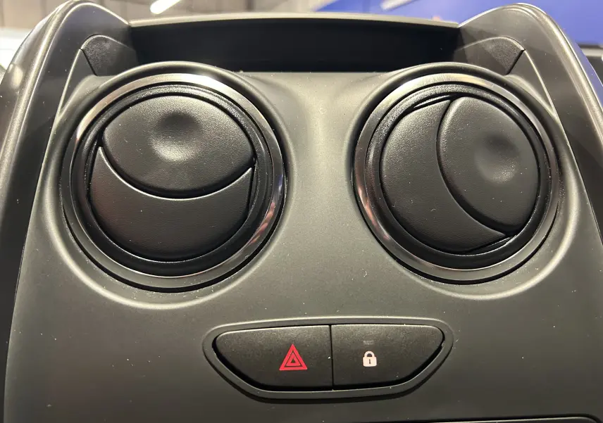 Gros plan sur les deux aérateurs ronds noirs et les boutons de verrouillage et warning du tableau de bord d'un Dacia Duster gris.