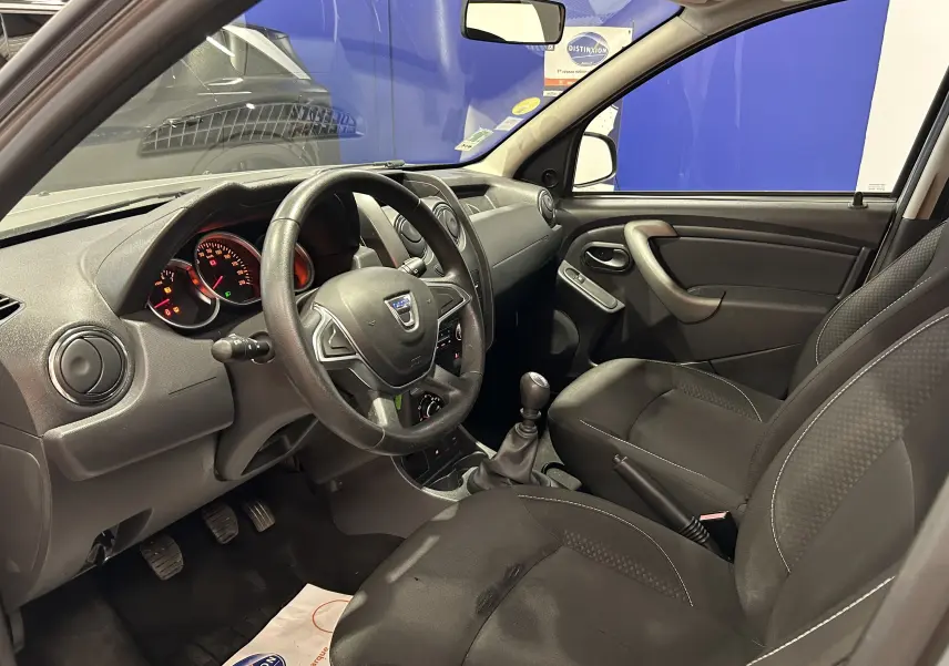 Intérieur avant droit du Dacia Duster gris 2017, volant cuir, tableau de bord simple et sièges tissu noirs.