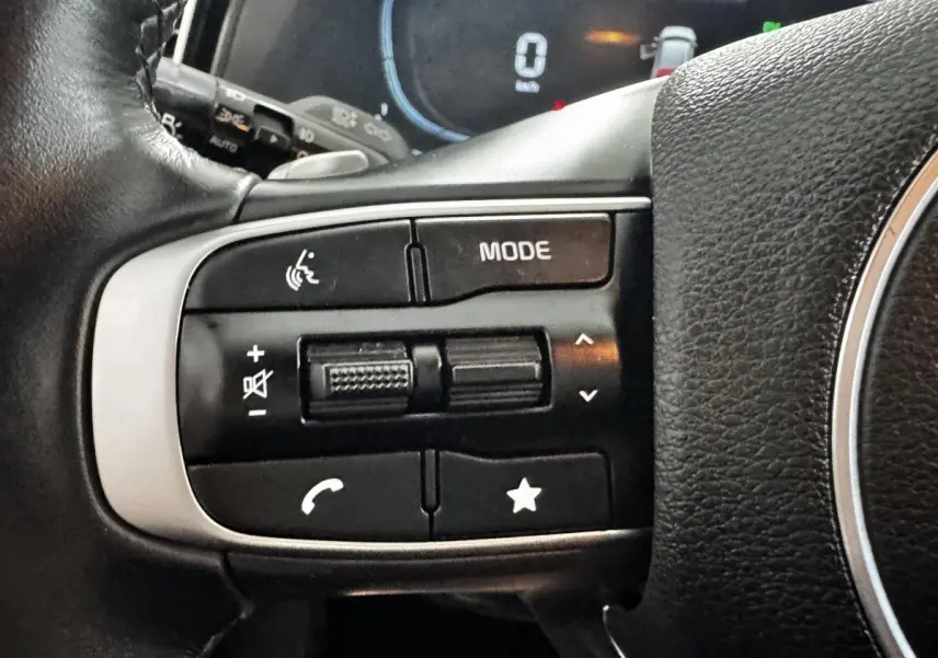 Gros plan sur les commandes gauche du volant cuir noir du Kia Sportage blanc 2024, avec boutons multifonctions.