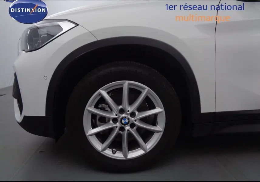 Gros plan sur la roue avant gauche d’un BMW X1 blanc Alpine Metal, mettant en valeur la jante en alliage et le phare avant.