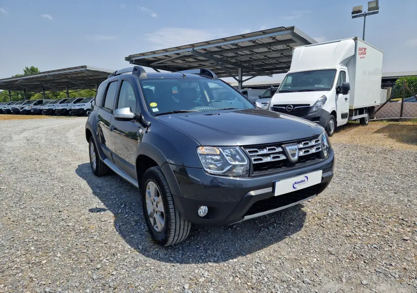 Vue 3/4 avant d'un Dacia Duster gris foncé métal 2015, avec calandre distinctive et barres de toit visibles.