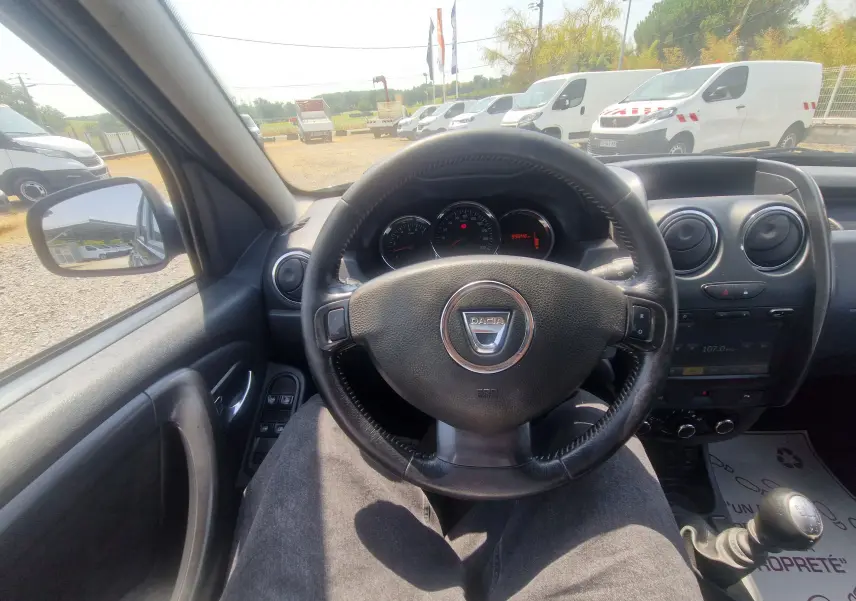 Vue intérieure centrée sur le volant noir du Dacia Duster 2015, avec tableau de bord et levier de vitesse manuelle visibles.