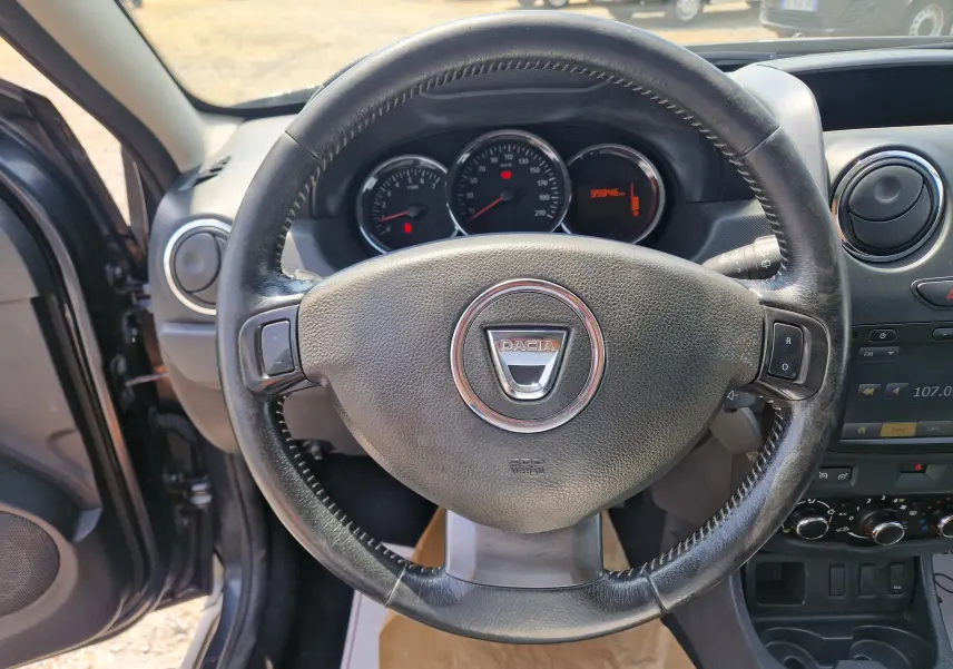 Vue intérieure centrée sur le volant en cuir noir du Dacia Duster gris foncé, avec tableau de bord et compteurs visibles.