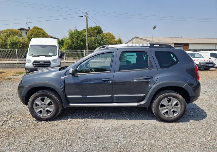 Vue de profil côté gauche d'un Dacia Duster gris foncé métal 2015 avec barres de toit et jantes alliage.