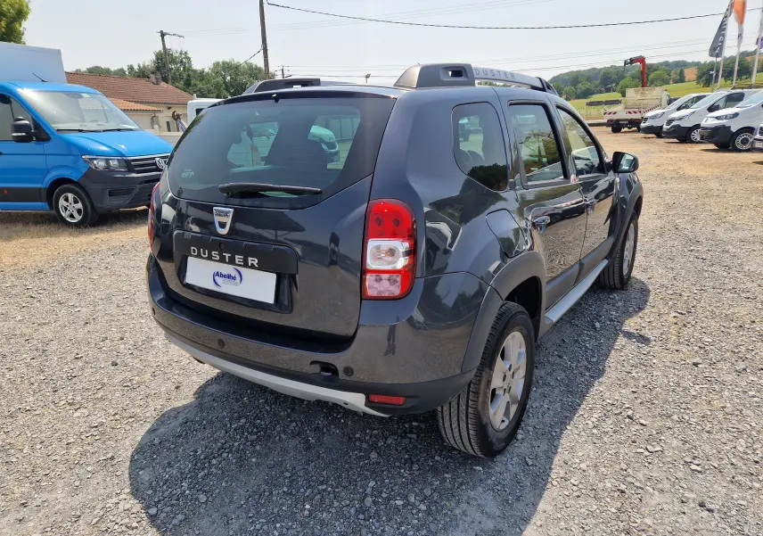 Vue 3/4 arrière droite d'un Dacia Duster gris foncé métal 2015, avec barres de toit et jantes alliage visibles.
