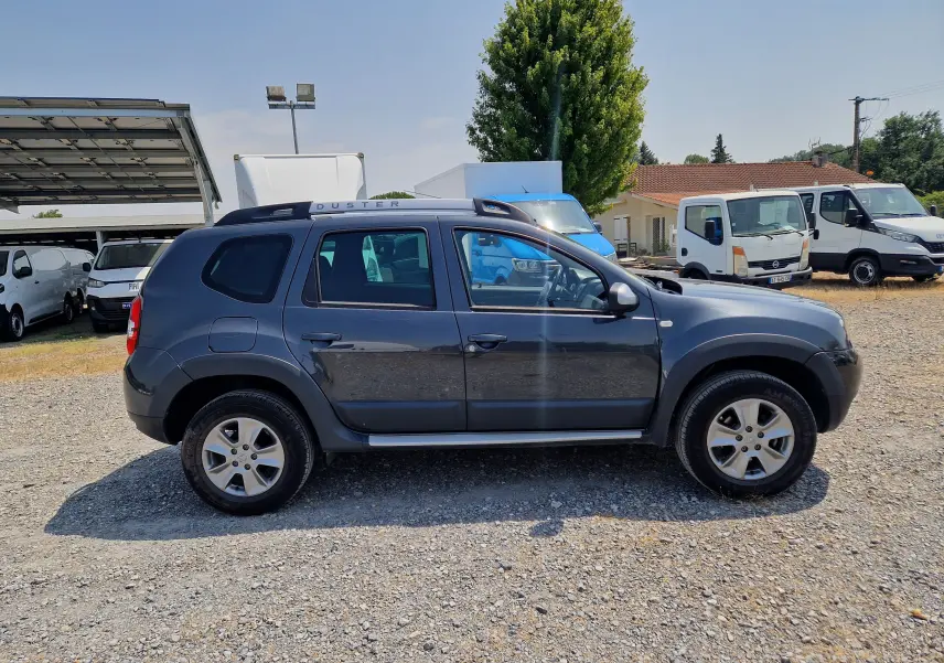 Profil côté droit d'un Dacia Duster gris foncé métal de 2015, avec barres de toit et jantes alliage visibles.