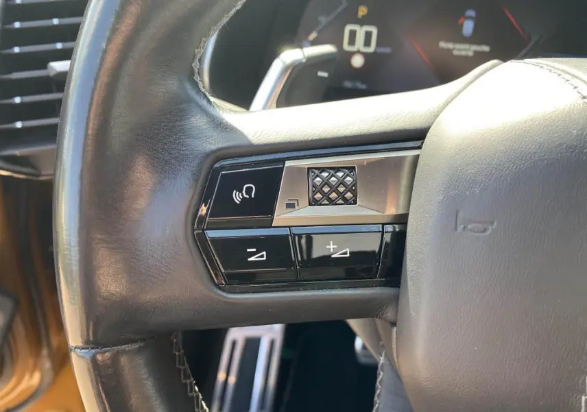 Gros plan sur les commandes gauche du volant cuir noir du DS7 Crossback jaune clair, avec tableau de bord numérique en arrière-plan.