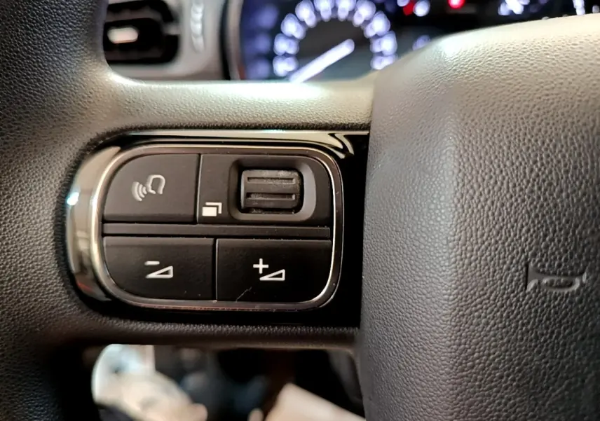 Gros plan sur les commandes de volume et téléphone du volant noir de la Citroën C3 blanche, tableau de bord flou en arrière-plan.